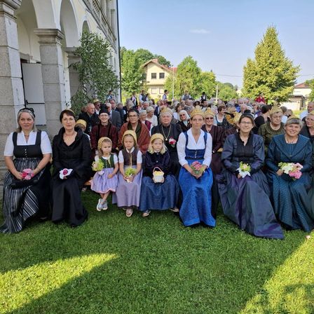Eine große Gruppe von Menschen, darunter Frauen und Kinder, versammelt sich auf einem Rasen vor einem Gebäude und hält Blumen in den Händen.