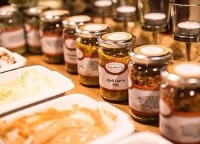 Ein Holztisch ist mit Gläsern verschiedener Pickles bedeckt, darunter eines mit der Aufschrift 'Kohl Curry Dip', und mehrere Teller mit Essen.