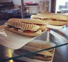 Eine Theke präsentiert vier Sandwiches auf weißen Tellern, mit getoastetem Brot und verschiedenen Füllungen. Sie stehen auf einem Glasregal über einem Tresen, wobei Küchengeräte im Hintergrund zu sehen sind.