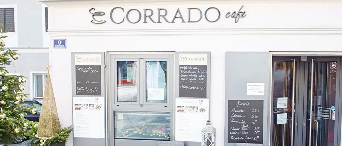 Das Äußere des Corrado Cafes in Österreich zeigt einen grauen Eingang mit Glastüren, Menütafeln und zwei Stühlen bei einem Tisch mit einer Laterne.