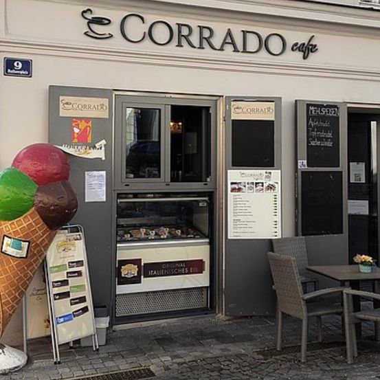 Corrado Cafe hat eine große Eisbecher-Statue vor der Tür. Ein Menü ist auf der Glastür sichtbar. Es gibt Outdoor-Stühle und Tische mit einer Blumenvase.