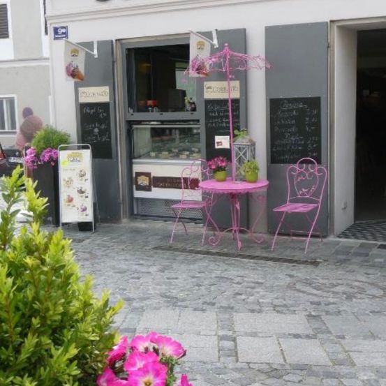 Ein Outdoor-Café mit pinken Tischen und Stühlern, einem Regenschirm und Blumen vor einem Gebäude mit einer Speisekarte und einer Person, die außerhalb steht.