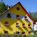 Ein leuchtend gelbes und weißes Haus mit rotem Rand und Blumenkästen, vor dem Hintergrund grüner Bäume und Berge.
