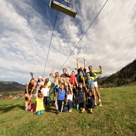 Eine Gruppe von Menschen posiert für ein Foto auf einer Wiese mit Bergen und einer Skilift-Anlage im Hintergrund.