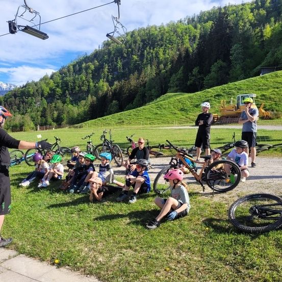 Auf einem Rasen sitzen Kinder mit Helmen auf dem Boden. In der Nähe sind Fahrräder abgestellt. Ein Mann steht und ein anderer hinter den Kindern. Ein Baufahrzeug ist auf der Straße geparkt. Bäume und Berge sind in der Ferne.