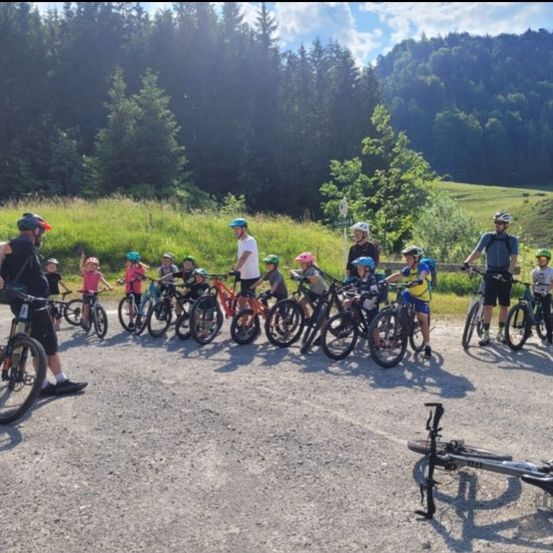 Eine Gruppe von Radfahrern, darunter Kinder, steht auf einem Kiesweg, umgeben von Grün und Bergen. Alle tragen Helme, mit Erwachsenen vorne und hinten.