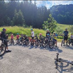 Eine Gruppe von Radfahrern, darunter Kinder, steht auf einem Kiesweg, umgeben von Grün und Bergen. Alle tragen Helme, mit Erwachsenen vorne und hinten.