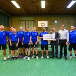 Eine Gruppe von Männern in blauen Shirts und Shorts steht in einem Fitnessstudio und posiert für ein Foto mit einem großen Scheck.