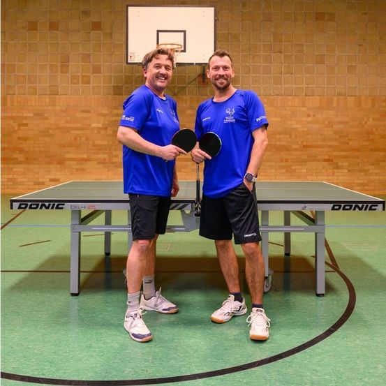 Zwei Männer in blauen T-Shirts stehen neben einem Tischtennistisch mit Schlägern, lächeln und posieren für ein Foto in einer Sporteinrichtung.