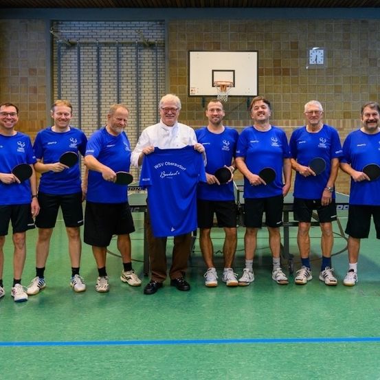 Eine Gruppe von Männern in blauen Shirts und schwarzen Shorts posiert für ein Foto und hält Tischtennisschläger. Ein Mann in einem weißen Hemd steht hinter ihnen und hält ein blaues Hemd. Eine Basketballkorb ist an der Wand hinter ihnen.