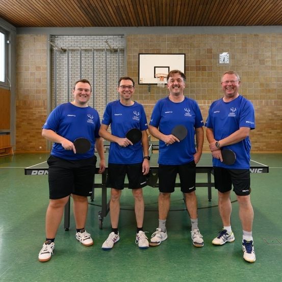 Vier Männer in blauen Shirts und schwarzen Shorts stehen mit Tischtennisschlägern in einer Turnhalle und lächeln. Vor ihnen steht ein Tischtennistisch. Dahinter ist ein Basketballkorb an der Wand montiert.