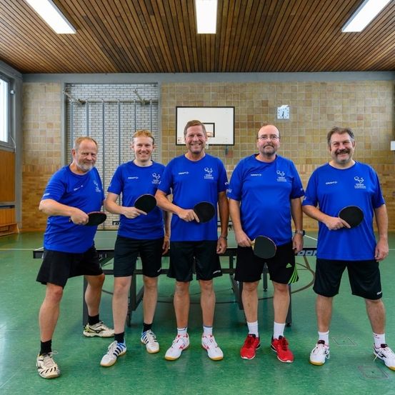 Fünf Männer in blauen T-Shirts und schwarzen Shorts stehen vor einem Tischtennistisch. Sie halten alle Schläger in den Händen und lächeln für die Kamera. Der Raum hat eine Holzdecke und Ziegelwände.