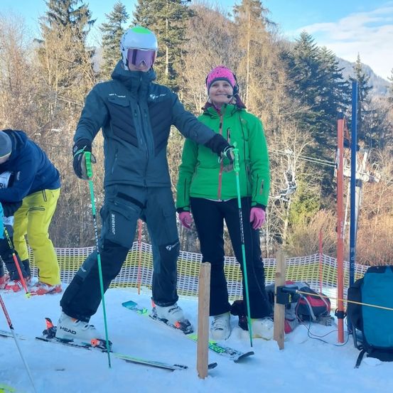 Zwei Skifahrer stehen auf einem verschneiten Hang. Der Mann trägt eine blaue Jacke und Helm, die Frau eine grüne Jacke und einen rosa Hut. Sie halten Skistöcke, und eine Seilbahn ist hinter ihnen.