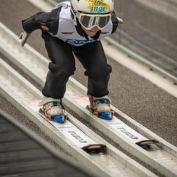 Ein Skifahrer in einem weißen Helm und mit Schutzbrille gleitet einen Skisprung hinunter, trägt einen Skianzug mit Inge auf dem Helm.