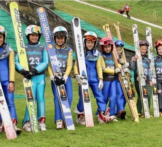 Eine Gruppe junger Skifahrer in passenden Uniformen posiert mit ihren Skiern für ein Foto vor einem Grasfeld.