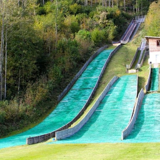 Ein langer, grün beschichteter Skisprung, umgeben von Bäumen und Gras. Ein kleines Holzgebäude ist in der Nähe.