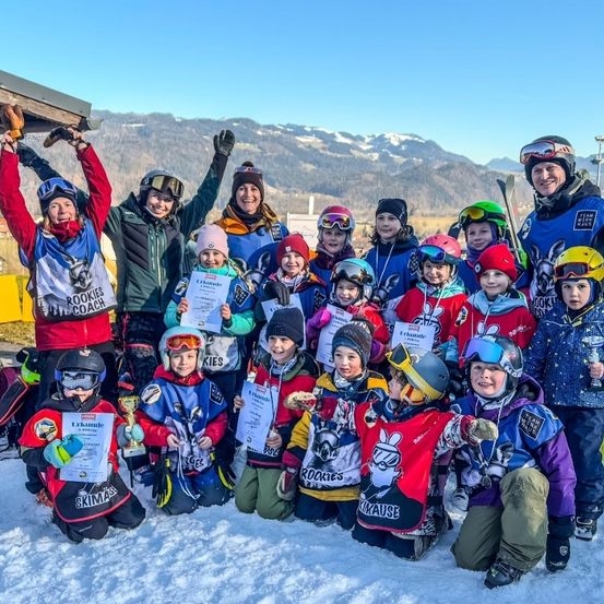 Eine Gruppe von Kindern und Erwachsenen in Skiausrüstung posiert für ein Foto auf einem verschneiten Berg. Einige halten Urkunden und Trophäen. Die Berge sind im Hintergrund.
