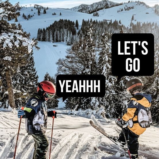 Zwei junge Skifahrer auf einem verschneiten Hang. Einer fährt bergab, während der andere steht und Skistöcke hält. Im Hintergrund befindet sich ein verschneiter Berg mit Tannen.