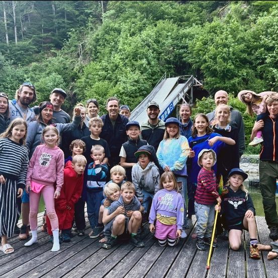 Eine Gruppe von Menschen, einschließlich Erwachsener und Kindern, versammelt sich auf einem hölzernen Steg in der Nähe eines Gewässers. Sie alle lächeln und posieren für ein Foto. Im Hintergrund sind eine Holzstruktur, eine Böschung und dichte grüne Bäume zu sehen.