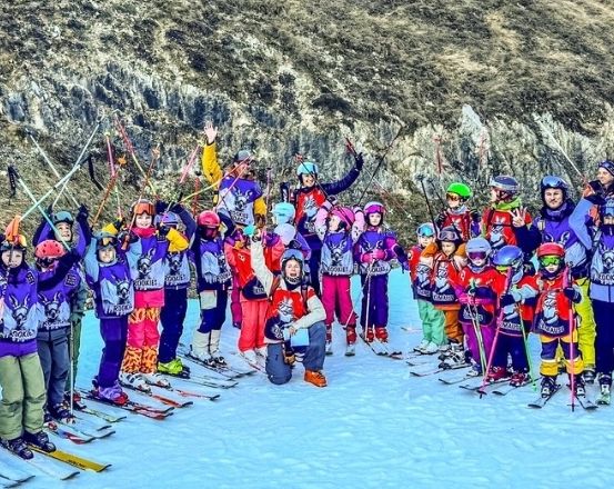 Eine Gruppe von Kindern und Erwachsenen posiert für ein Foto auf verschneiten Hängen mit Skiausrüstung, einige mit Helmen, vor einem felsigen Berg.
