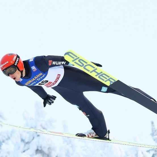 Ein Skifahrer in einem blauen und roten Skianzug mit der Nummer 45 schwebt in der Luft auf FISCHER-markierten Skiern vor einem verschneiten Hintergrund.