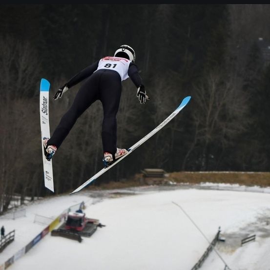 Ein Skifahrer in einem schwarzen Skianzug springt in die Luft, trägt einen Helm, Handschuhe und Skischuhe. Der Skifahrer hält Skistöcke. Der Schnee ist weiß, und es gibt Bäume im Hintergrund.