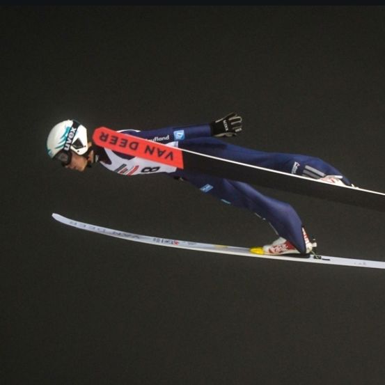 Ein Skifahrer befindet sich in der Luft mit weit auseinander gespreizten Skiern. Der Skifahrer trägt einen blauen Anzug und einen Helm. Das Wort 'VAV' ist auf dem Rücken des Skifahrers geschrieben.
