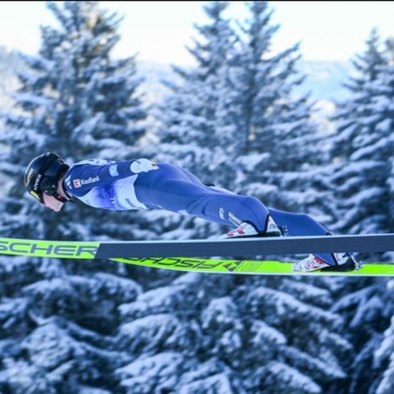 Ein Skispringer schwebt mit blau-weißer Uniform, Helm und Skiern in der Luft, umgeben von verschneiten Tannen.