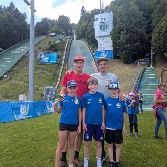 Drei Männer und drei Kinder stehen auf einem Rasenfeld in der Nähe einer Skisprungschanze. Die Kinder halten einen Pokal, und der Mann links trägt einen Hut.