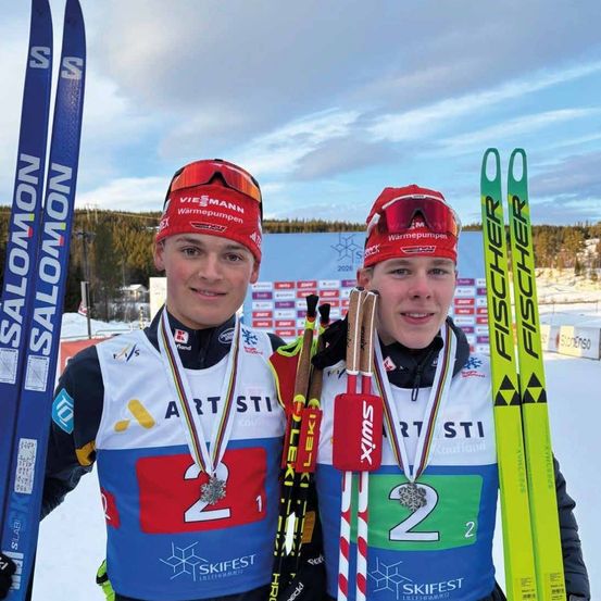 Zwei männliche Skifahrer mit roten Kopfbändern und Medaillen stehen gemeinsam auf einem verschneiten Feld, halten ihre Skistöcke und lächeln für die Kamera. Hinter ihnen befindet sich ein Hintergrund mit einem Schild, auf dem 'Warmpumpen' steht.
