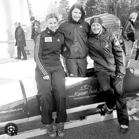 Drei Frauen in Winterjacken stehen neben einem Bobsled und lächeln. Die Frau links trägt eine Jacke mit weißen Streifen. Bäume und ein Gebäude sind im Hintergrund zu sehen.