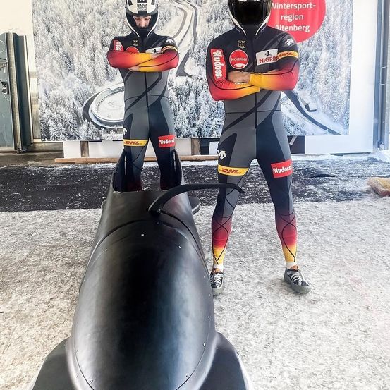 Zwei Männer in Bobsleigh-Ausrüstung stehen neben einem Bobsleigh mit einer Kulisse, die eine Wintersportregion zeigt. Beide tragen Helme und haben die Arme verschränkt.