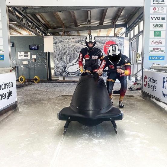 Zwei Athleten bereiten sich in einem Bobschlitten auf einer Wintersportanlage vor, tragen Helme und Rennanzüge. Hinter ihnen befindet sich eine große Wand mit einer schneebedeckten Landschaft und mehreren Werbeplakaten.
