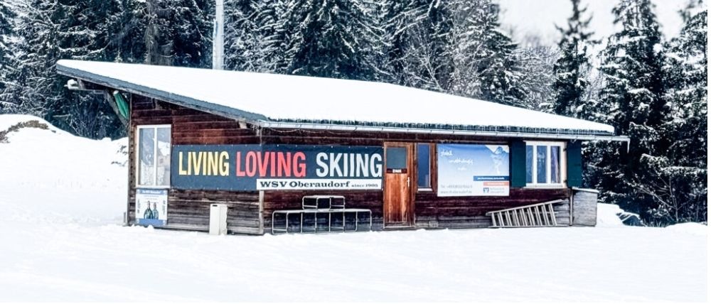 Ein schneiebedecktes Holzhäuschen mit einem blauen Schild, das 'Leben Lieben Skifahren' sagt. Bänke und eine Leiter sind draußen.