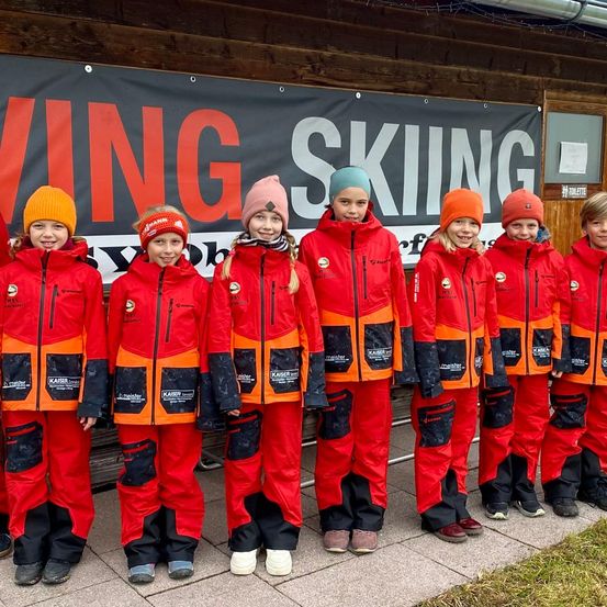 Eine Gruppe von Kindern in roten und orangefarbenen Skianzügen steht vor einem Gebäude mit einem Banner, das 'WING SKIING' besagt.