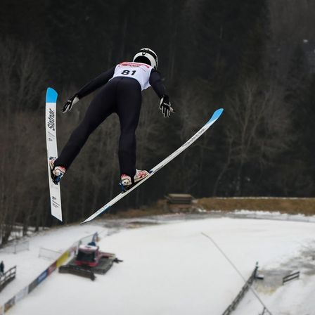 Ein Skifahrer in schwarzem und weißem Outfit und Helm springt in der Luft mit gespreizten Skiern auf einer verschneiten Piste.
