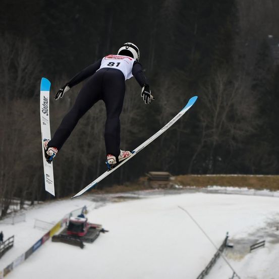 Ein Skifahrer in schwarzem und weißem Outfit und Helm springt in der Luft mit gespreizten Skiern auf einer verschneiten Piste.
