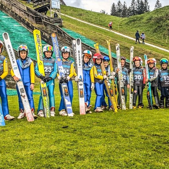 Eine Gruppe junger Skifahrer in passenden Uniformen steht mit ihren Skiern auf einem Grasfeld in der Nähe einer Skipiste. Sie tragen Helme und Handschuhe.