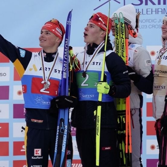 Vier Athleten stehen auf einem Podest und feiern mit Skistöcken. Zwei tragen rote Stirnbänder und alle tragen Medaillen. Sie stehen vor einer blauen Wand mit verschiedenen Sponsorenlogos.