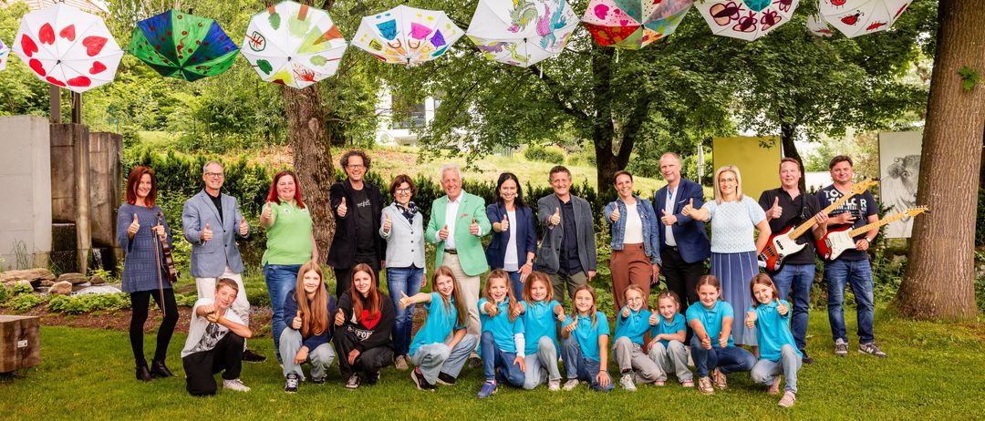 Eine Gruppe von Erwachsenen und Kindern posiert auf einem Rasen für ein Foto, unter dekorierten Regenschirmen, die von Bäumen hängen.