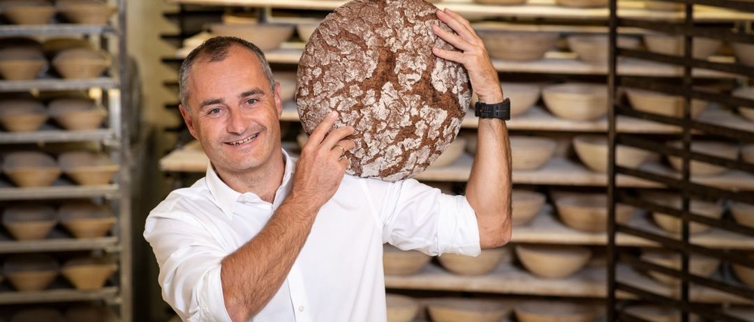Ein Mann hält ein großes Brot in einer Bäckerei, im Hintergrund befinden sich Regale mit Schüsseln.