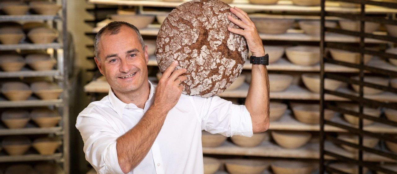 Ein Mann hält ein großes Brot in einer Bäckerei, im Hintergrund befinden sich Regale mit Schüsseln.