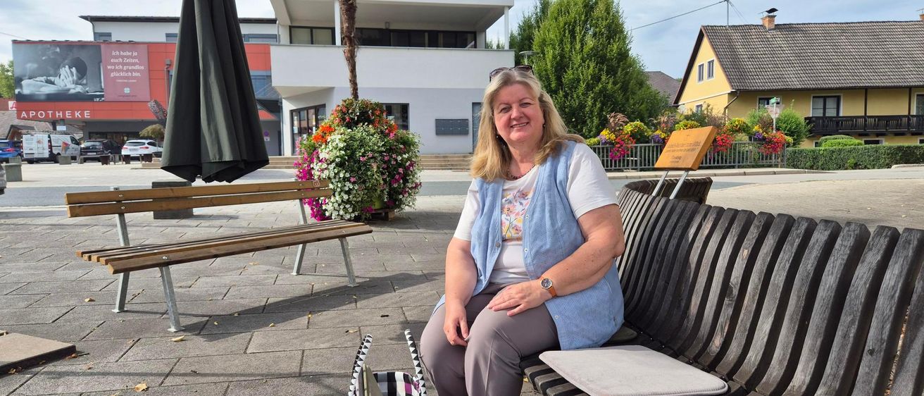 Eine Frau sitzt lächelnd auf einer Bank in einem Stadtplatz. Neben ihr steht eine schwarz-weiß karierte Tasche. Hinter ihr befindet sich ein Gebäude, ein Baum und ein Haus.