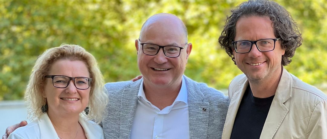 Drei Menschen stehen eng beieinander für ein Foto. Der Mann links trägt einen Blazer und eine Halskette. Der Mann in der Mitte trägt eine Brille. Die Frau rechts trägt einen weißen Blazer. Hinter ihnen steht ein grüner Busch.
