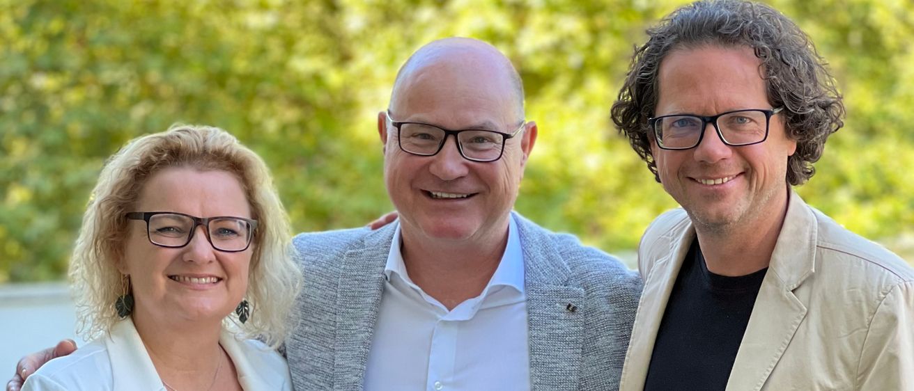 Drei Menschen stehen eng beieinander für ein Foto. Der Mann links trägt einen Blazer und eine Halskette. Der Mann in der Mitte trägt eine Brille. Die Frau rechts trägt einen weißen Blazer. Hinter ihnen steht ein grüner Busch.