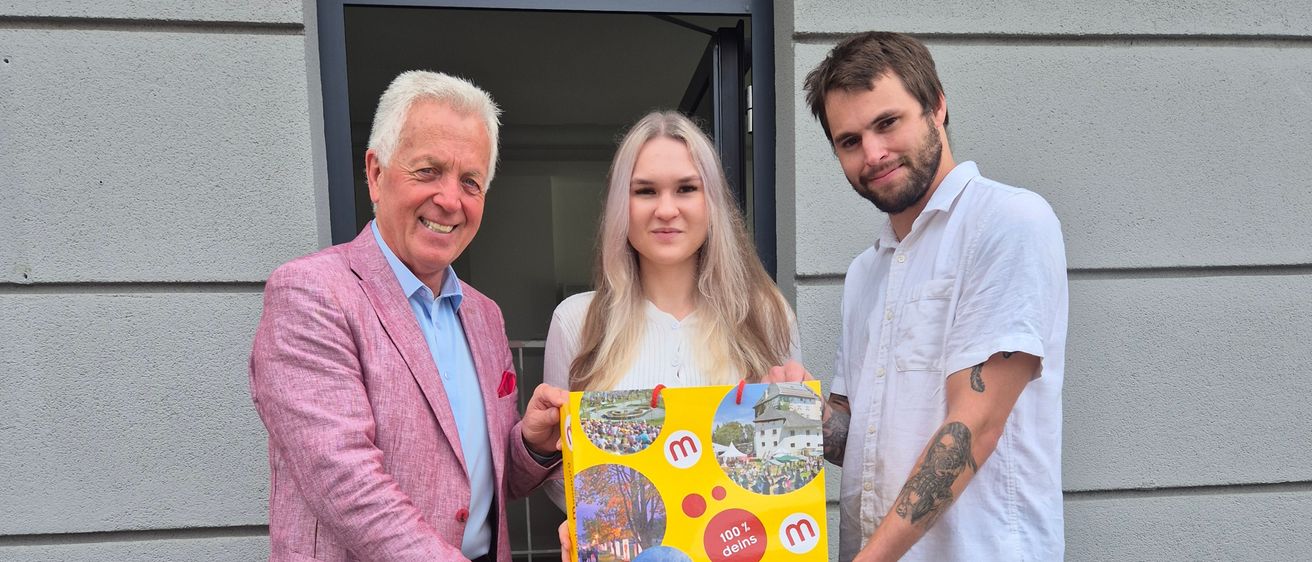 Drei Personen posieren für ein Bild und halten eine gelbe Box mit Bildern und Text. Der Mann auf der linken Seite trägt einen rosa Anzug, die Frau ein weißes Hemd und einen schwarzen Rock und der Mann auf der rechten Seite trägt ein weißes Hemd und Jeans. Sie stehen auf Betonstufen vor einem Gebäude.