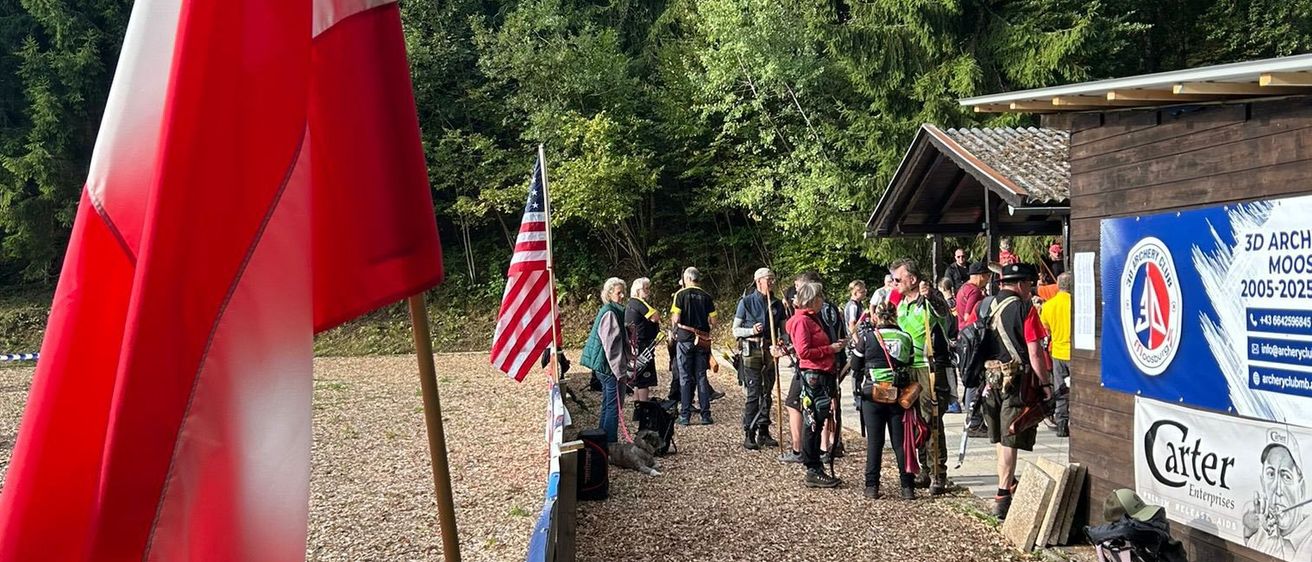 Eine Gruppe von Menschen versammelt sich in der Nähe einer Flagge und eines Gebäudes. Sie tragen Rucksäcke. Es gibt ein Holzgebäude mit einem Schild und einem Plakat an der Seite. Bäume sind im Hintergrund.