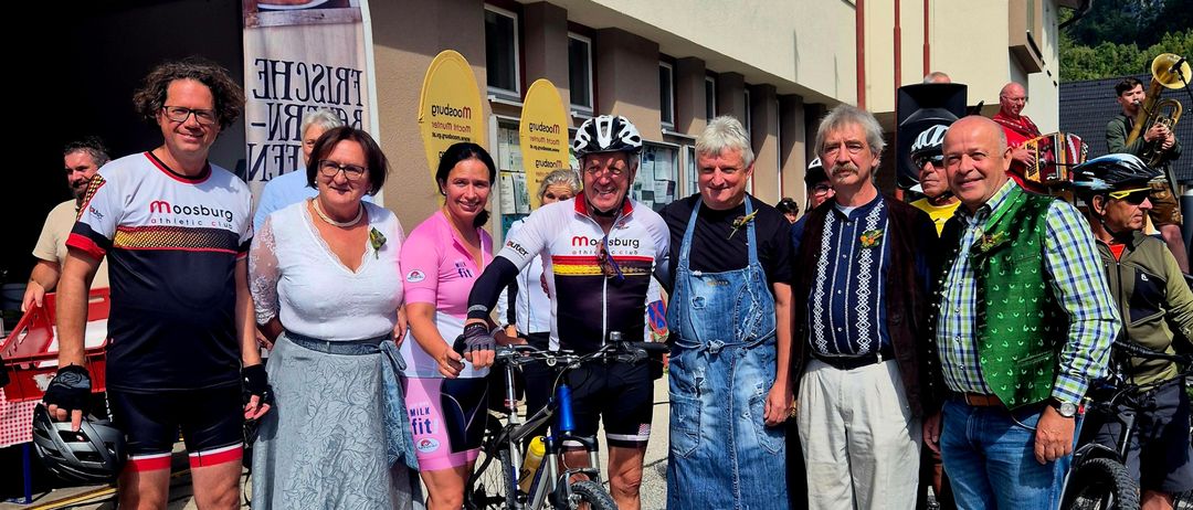 Eine Gruppe von Menschen steht vor einem Gebäude. Einige tragen Radsportbekleidung. Ein Mann hält ein Fahrrad, während andere Freizeitkleidung tragen. Musikinstrumente und eine Fahne sind in der Nähe.