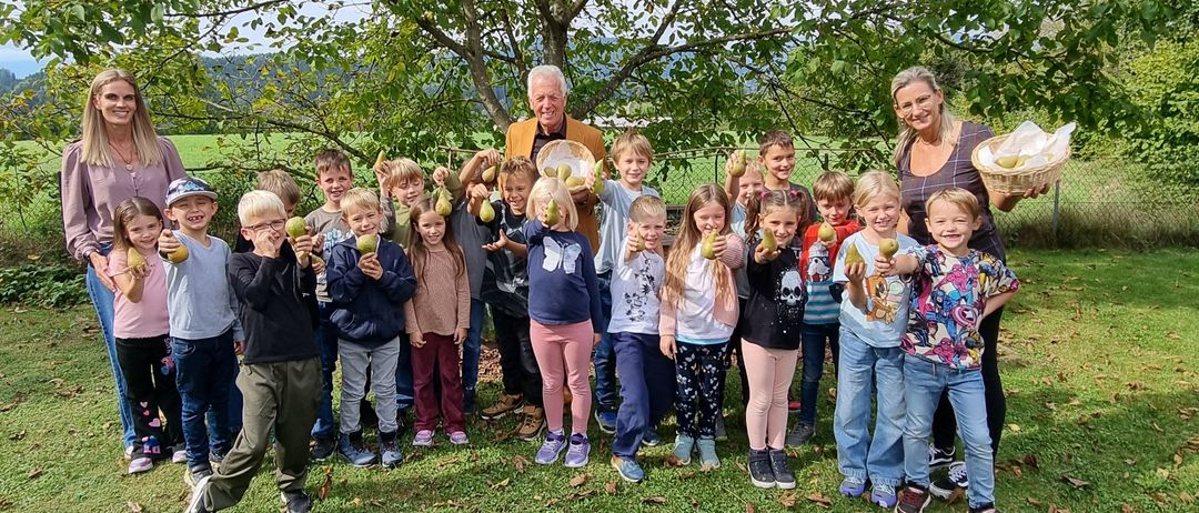 Eine Gruppe von Kindern und Erwachsenen steht vor einem Baum, hält Birnen und genießt einen sonnigen Tag.