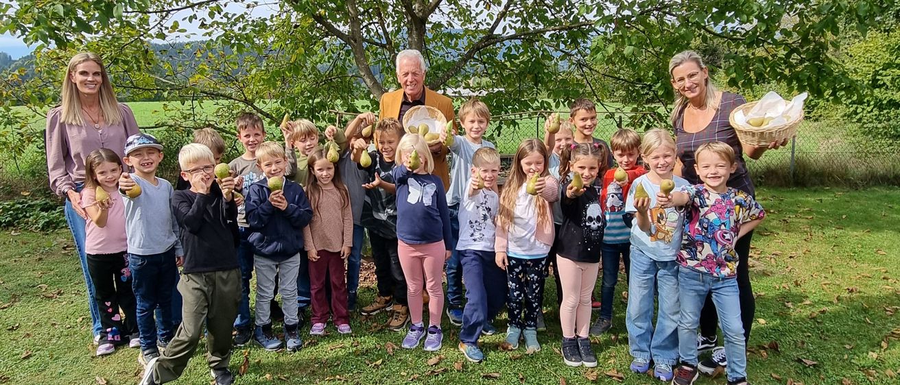Eine Gruppe von Kindern und Erwachsenen steht vor einem Baum, hält Birnen und genießt einen sonnigen Tag.